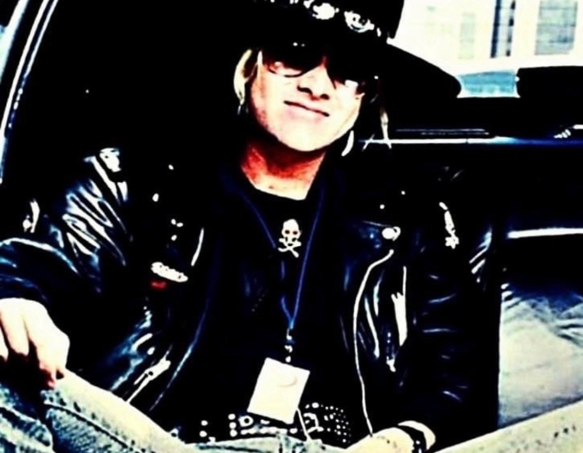 Mark Kendall in a black leather jacket, sunglasses and hat leaning on a car