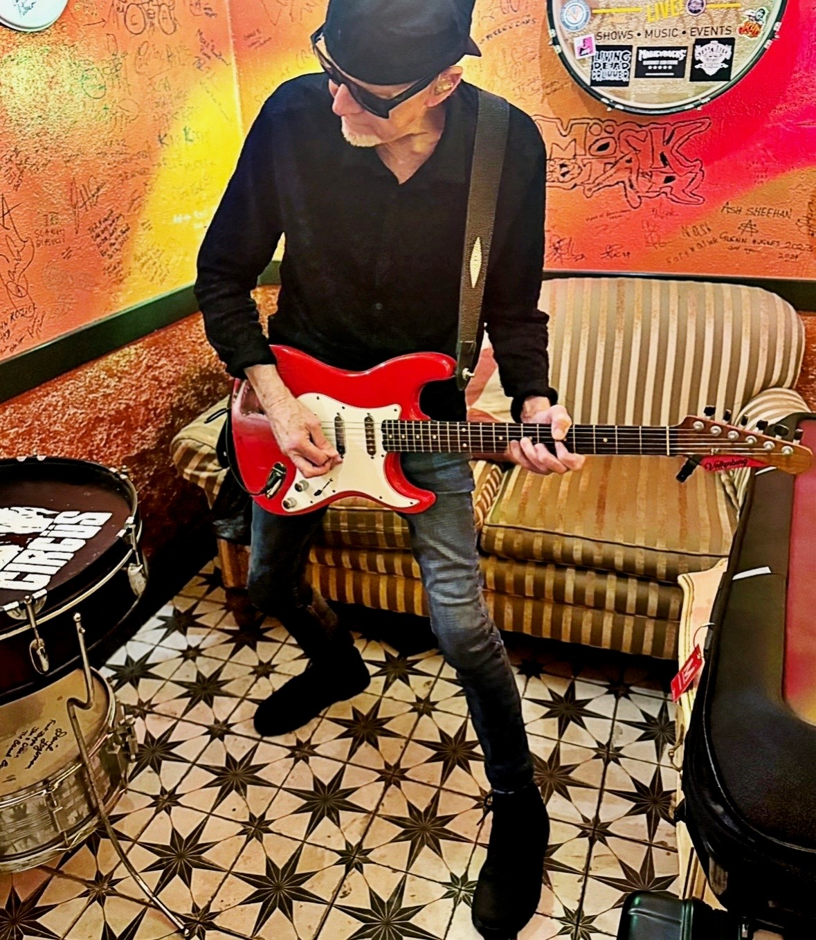 Mark Kendall playing a red electric guitar in a graffiti-covered green room