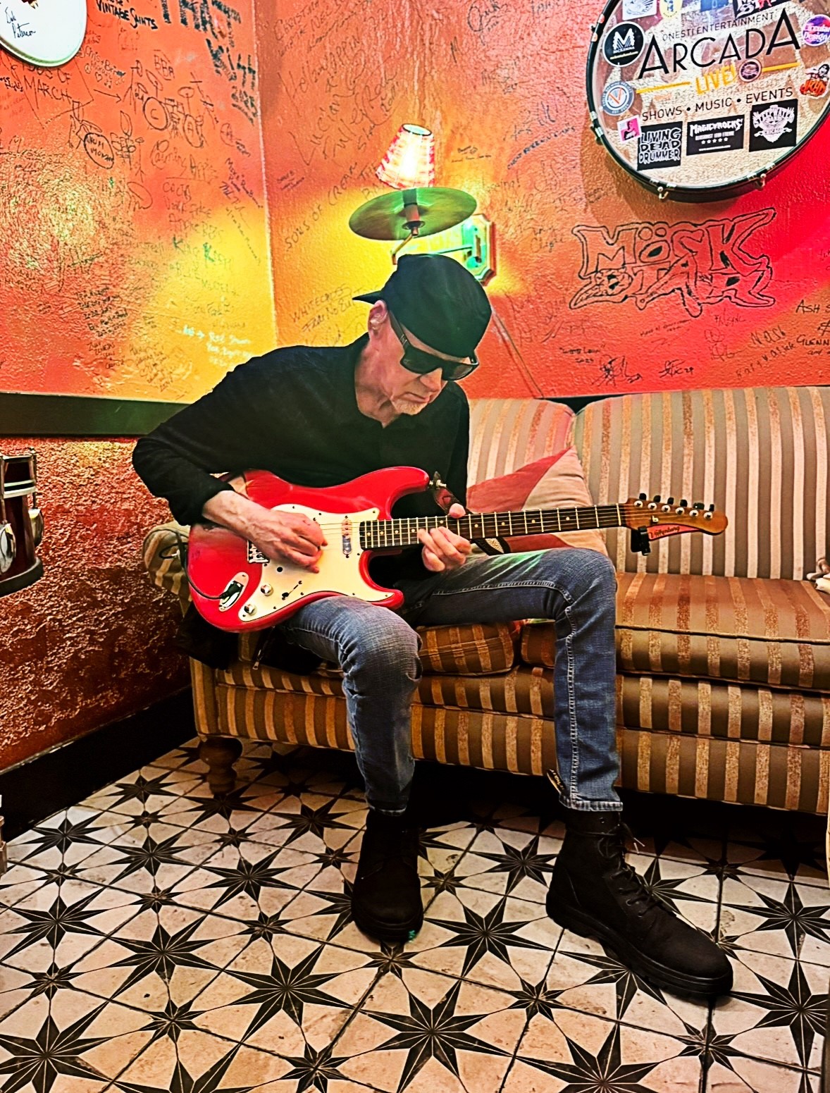 Mark Kendall seated backstage playing a red Stratocaster guitar